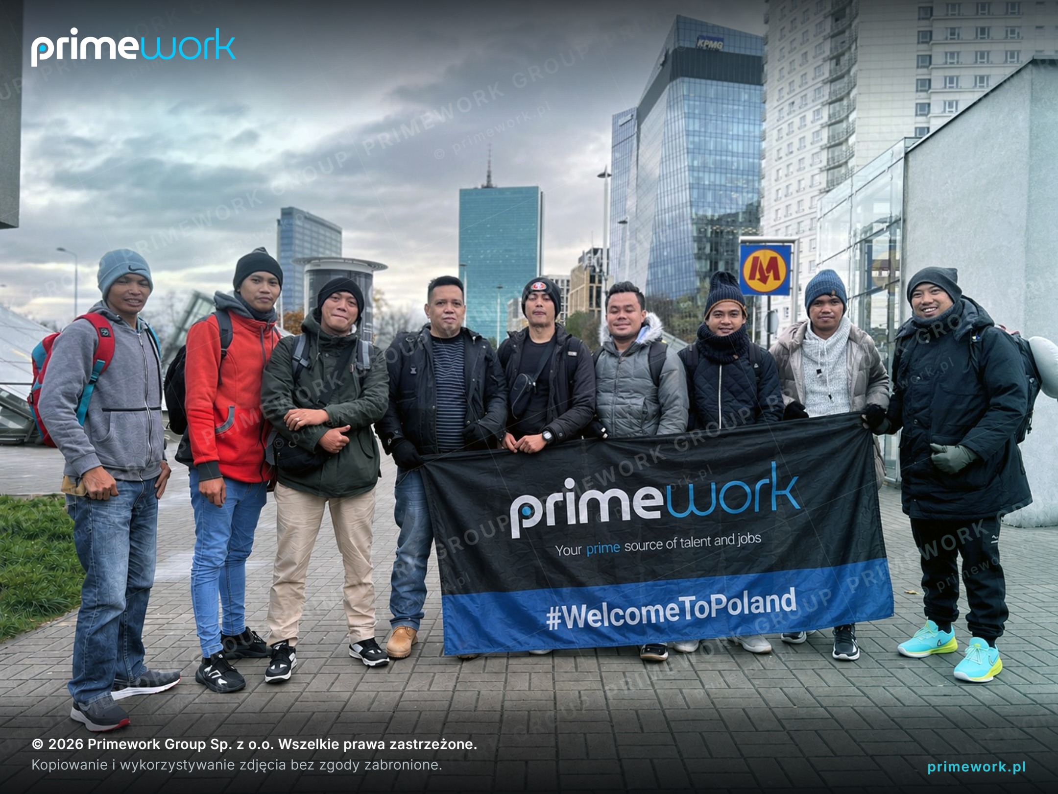 Grupa pracowników z Azji z banerem Primework #WelcomeToPoland w centrum Warszawy